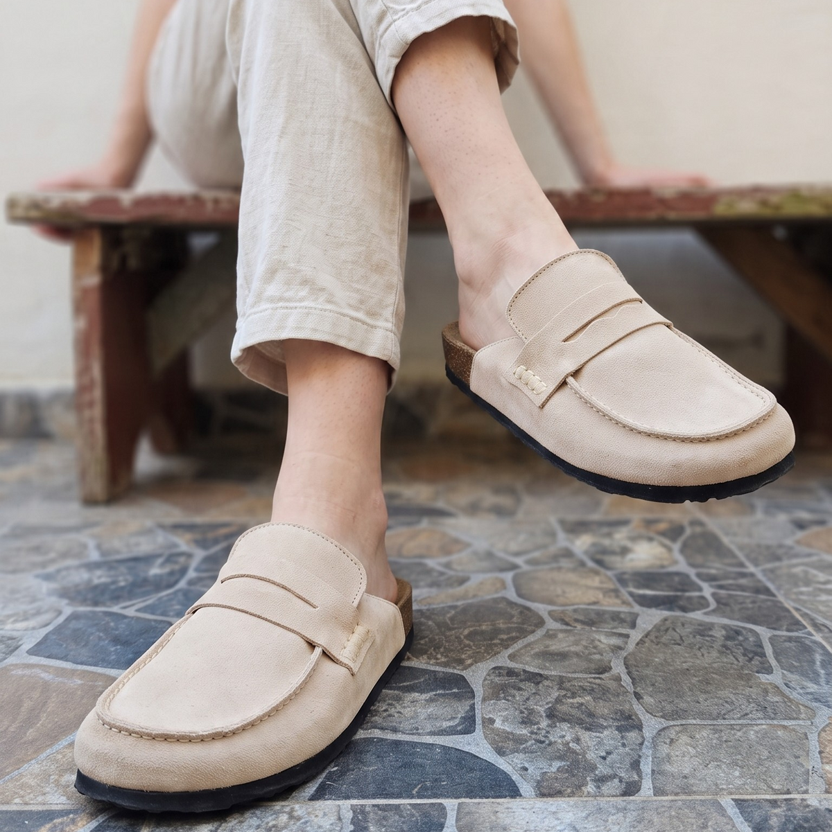 Mules Avern Dune en cuir beige, portées avec un pantalon écru sur un banc